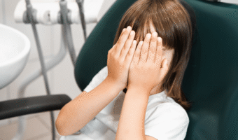 Child sitting in a dental chair, covering their eyes.
