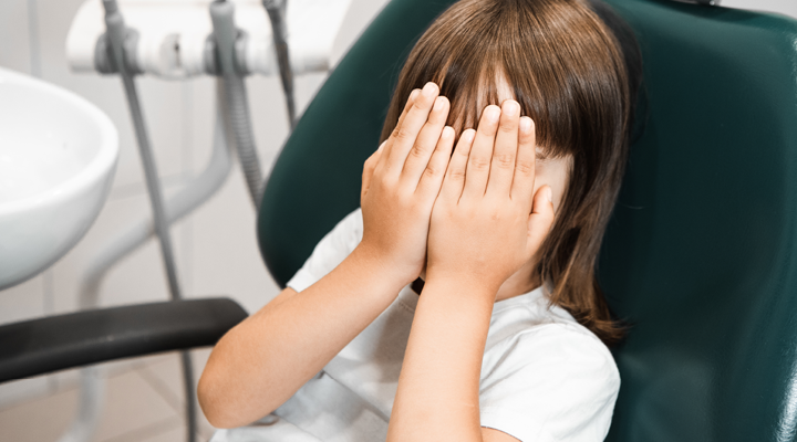 Child sitting in a dental chair, covering their eyes.