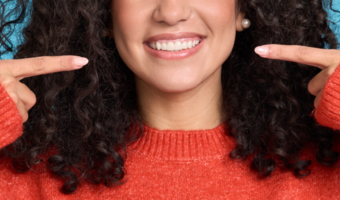 A woman in a red sweater points to her smiling mouth with box index fingers.