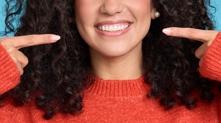 A woman in a red sweater is point toward her smiling mouth with both index fingers.